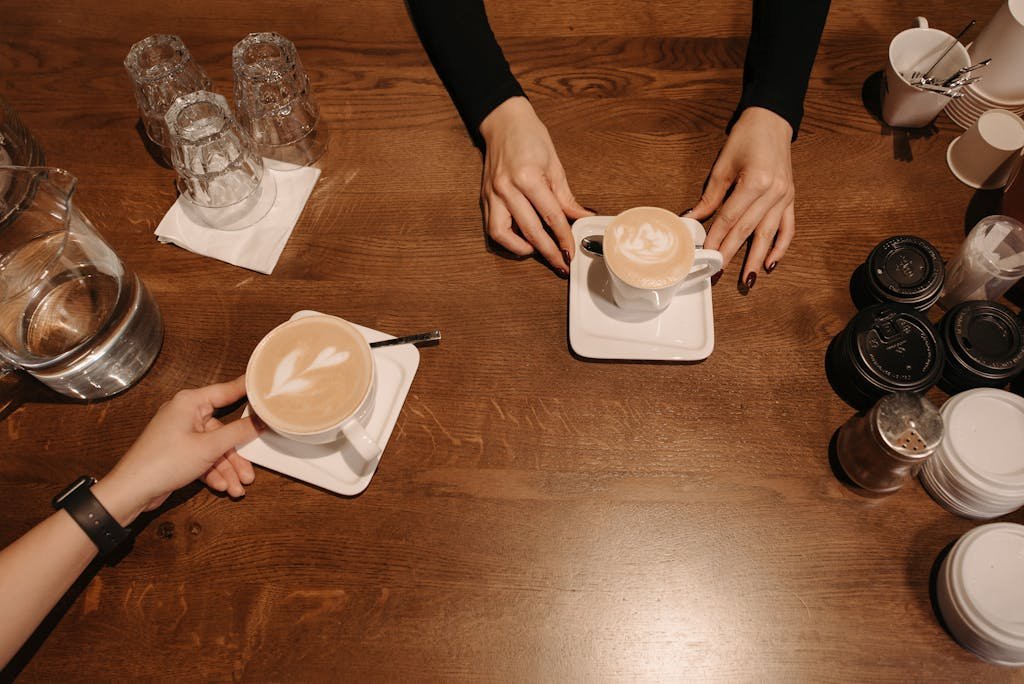 Two baristas serve lattes with foam art on a wooden table in a cozy coffee shop.